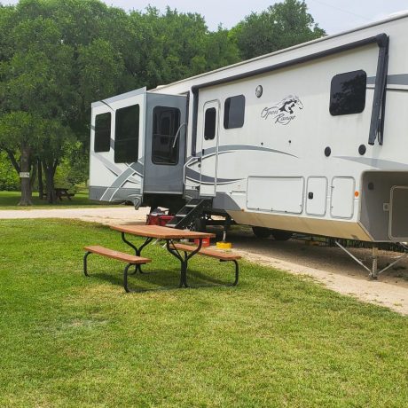 Large 5th wheel in a pull thru RV site with a grass yard and picnic table.