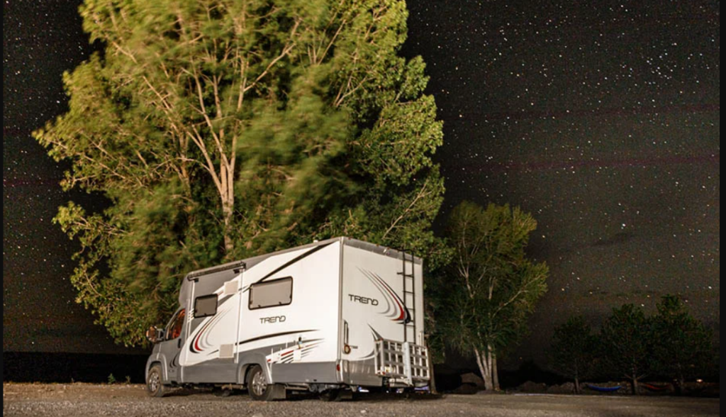 A class C Motorhome parked under a tree at night with a night sky full of stars.